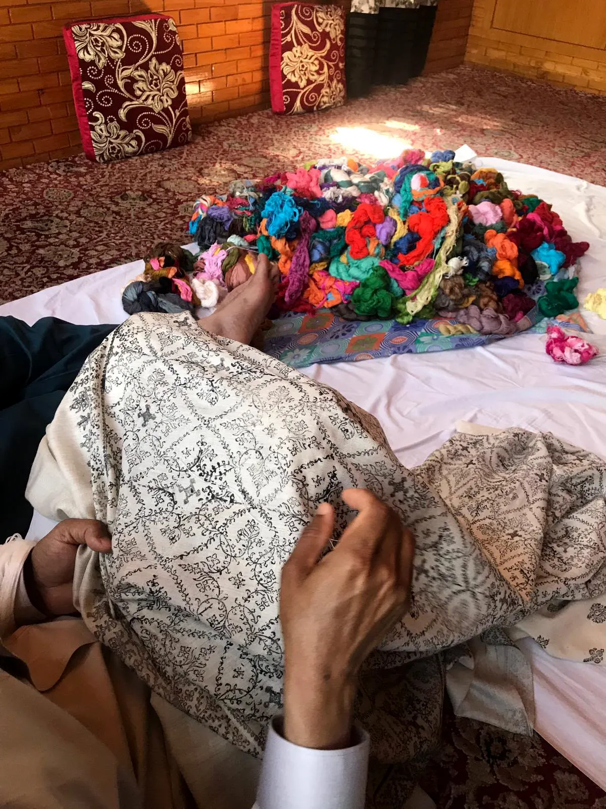 Hands pulling a thread through a printed pattern on an ivory shawl, a heap of coloured yarn behind.