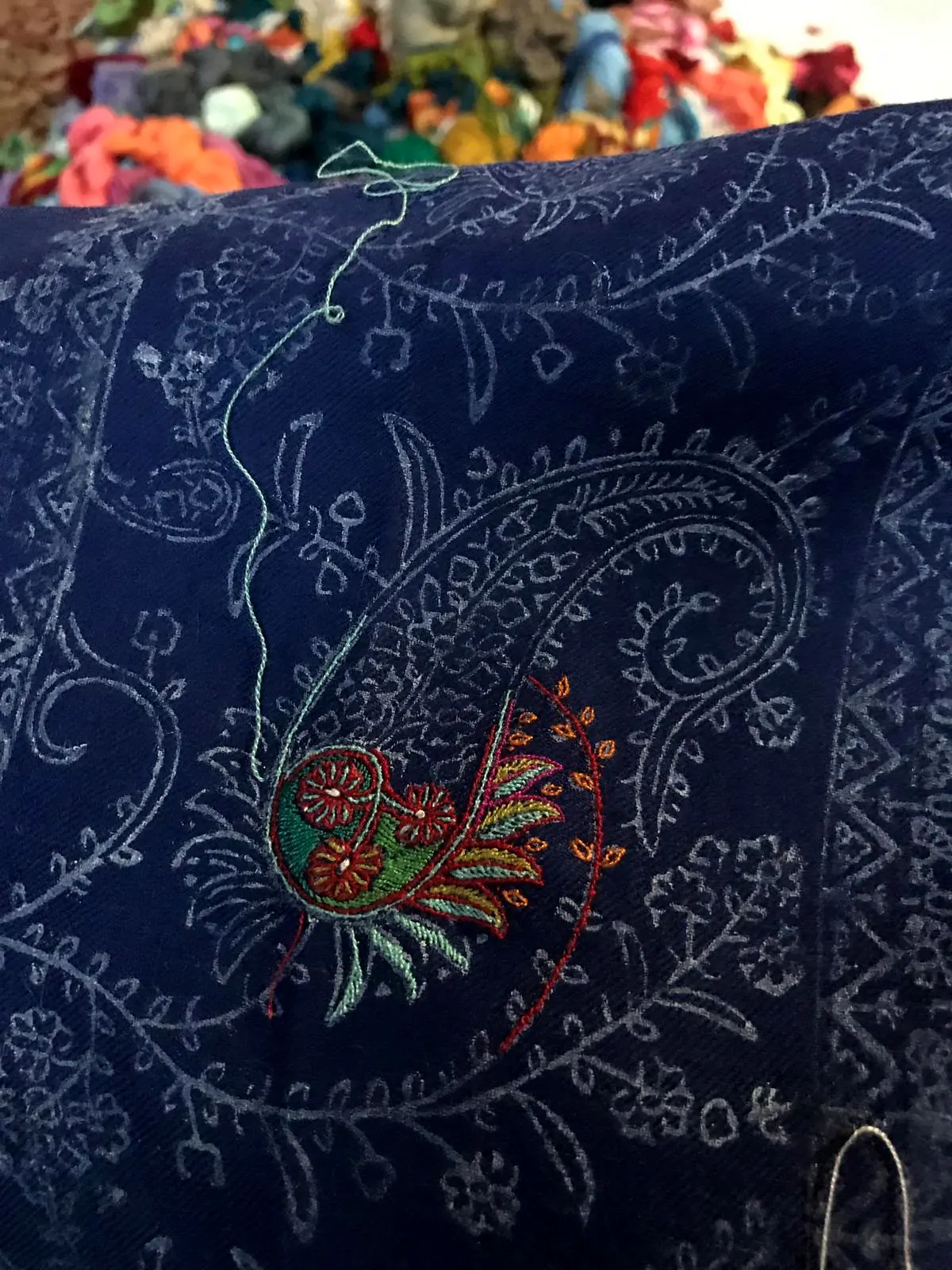 A navy shawl printed with a paisley outline, one corner already filled in with coloured needlework.