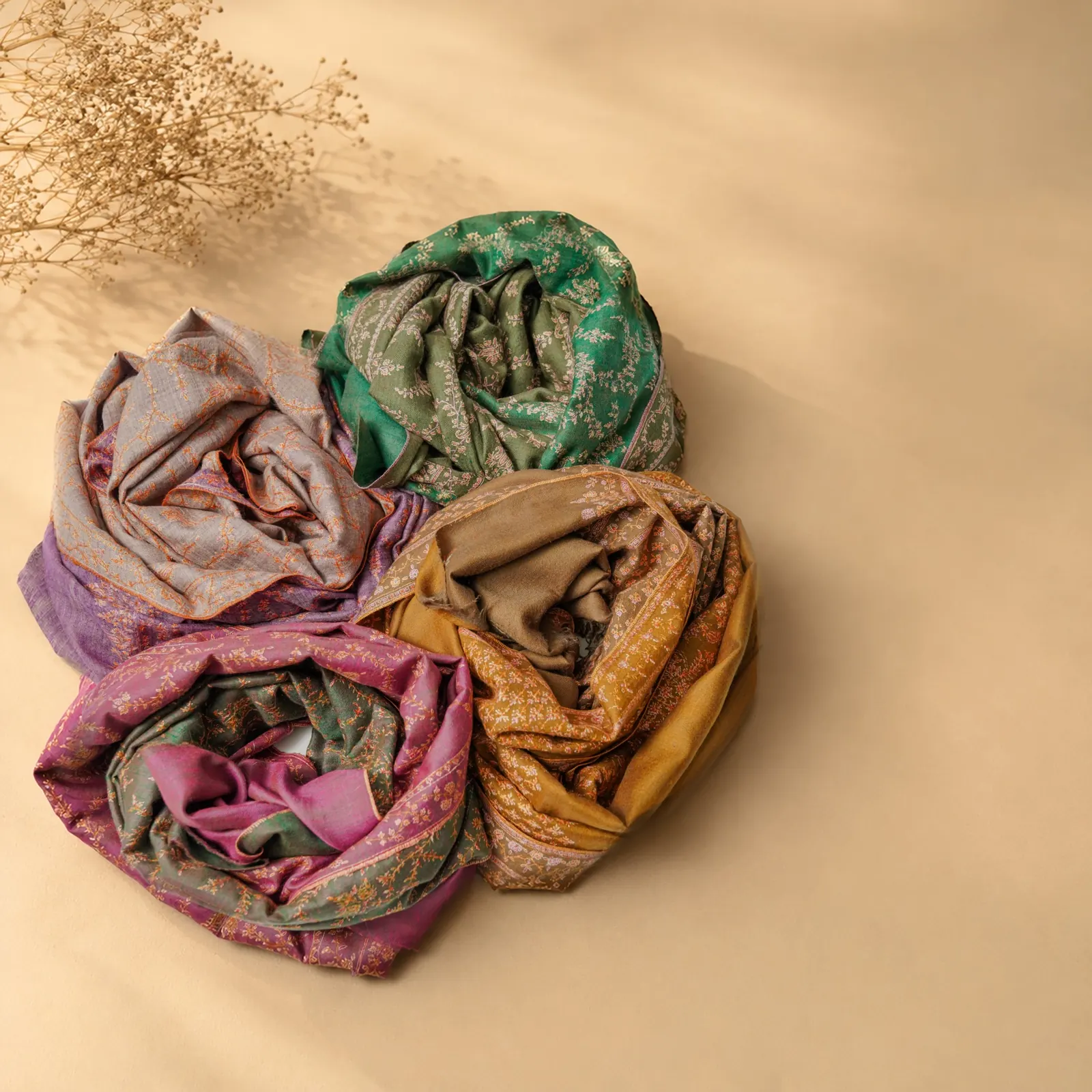 Four shawls rolled and arranged together in lilac, green, magenta and gold, on a sand ground.