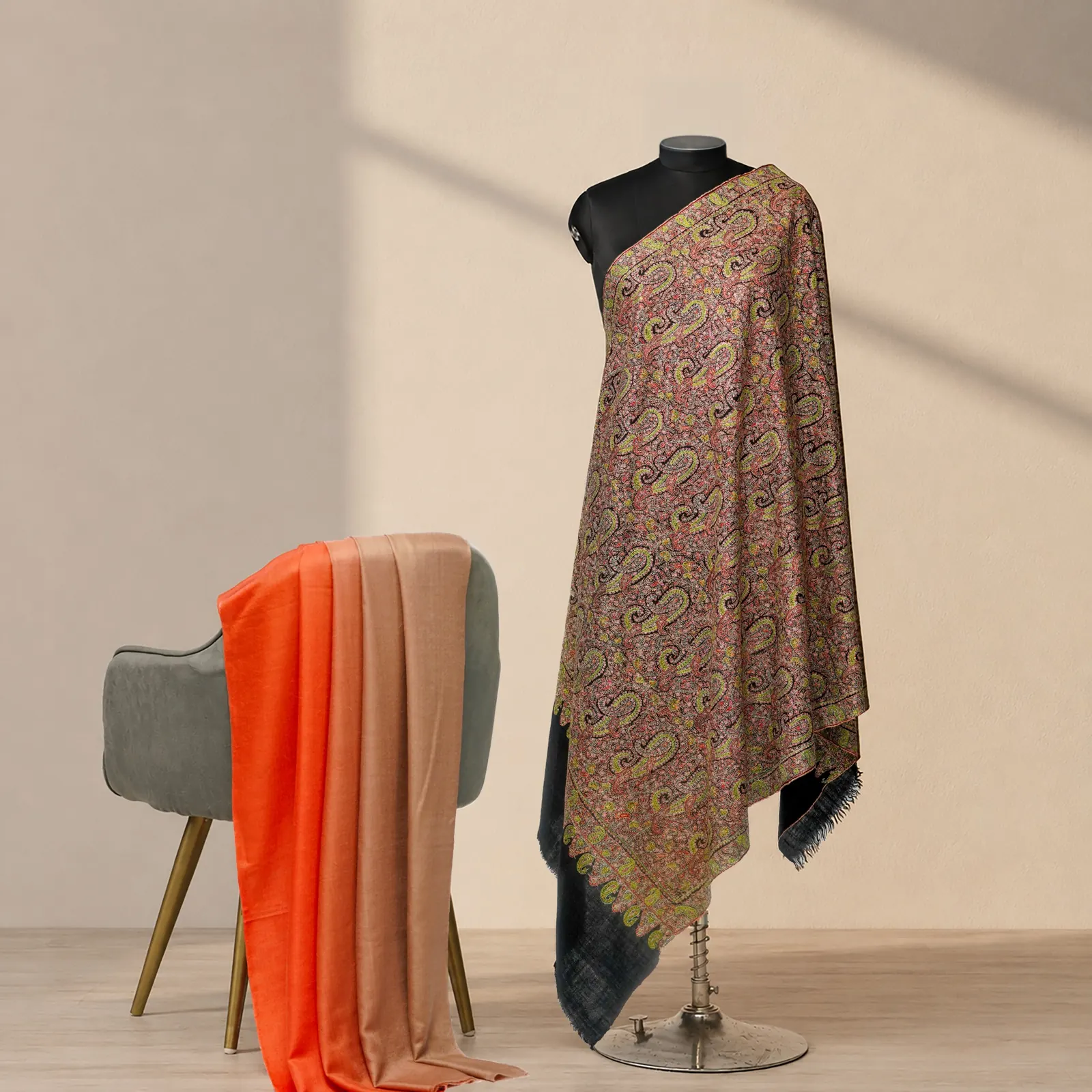 Shawl with fine paisley in rose, olive and violet on a stand, an orange shawl folded over a chair behind.