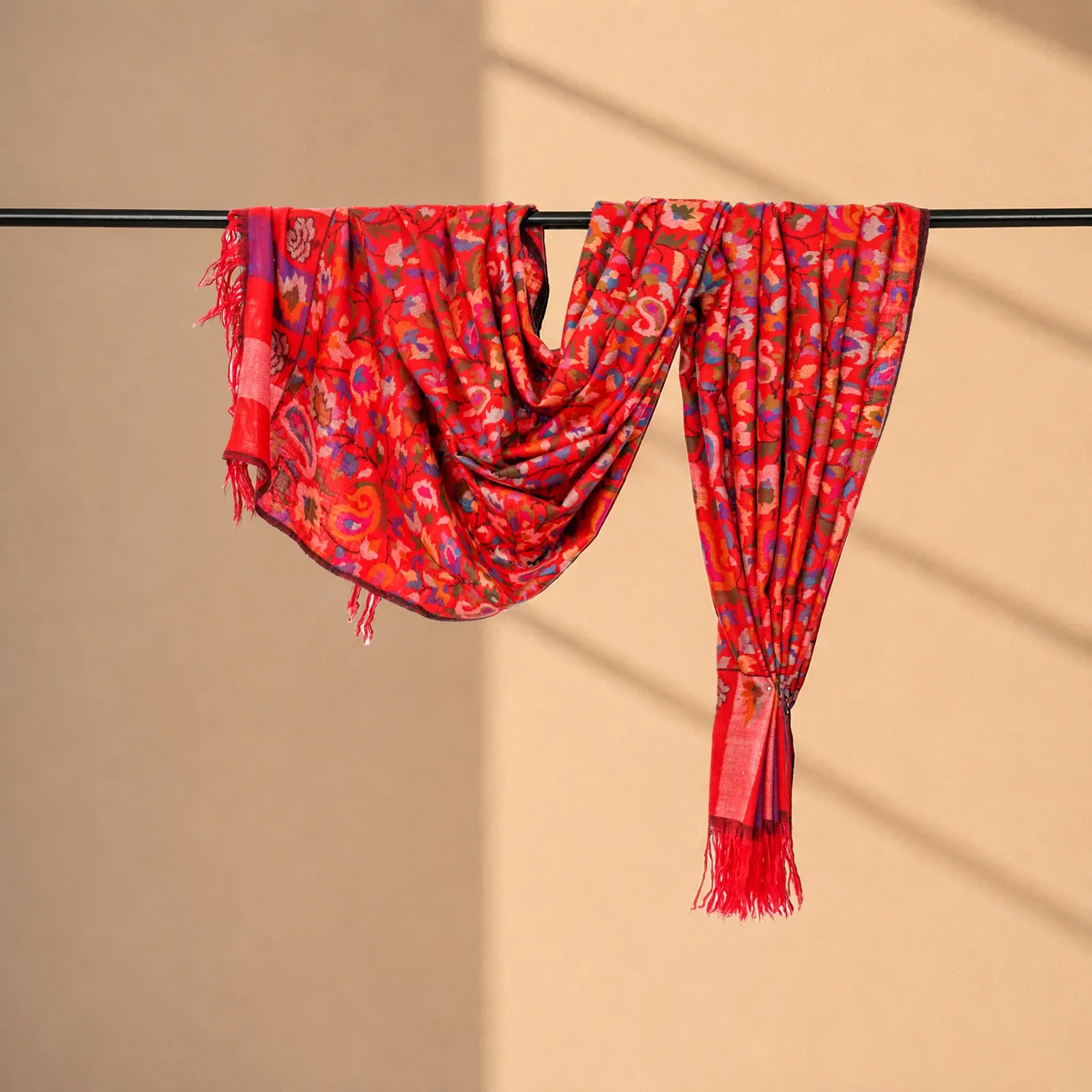 Scarlet shawl scattered with floral and paisley motifs in blue, violet and gold, hung from a rod.