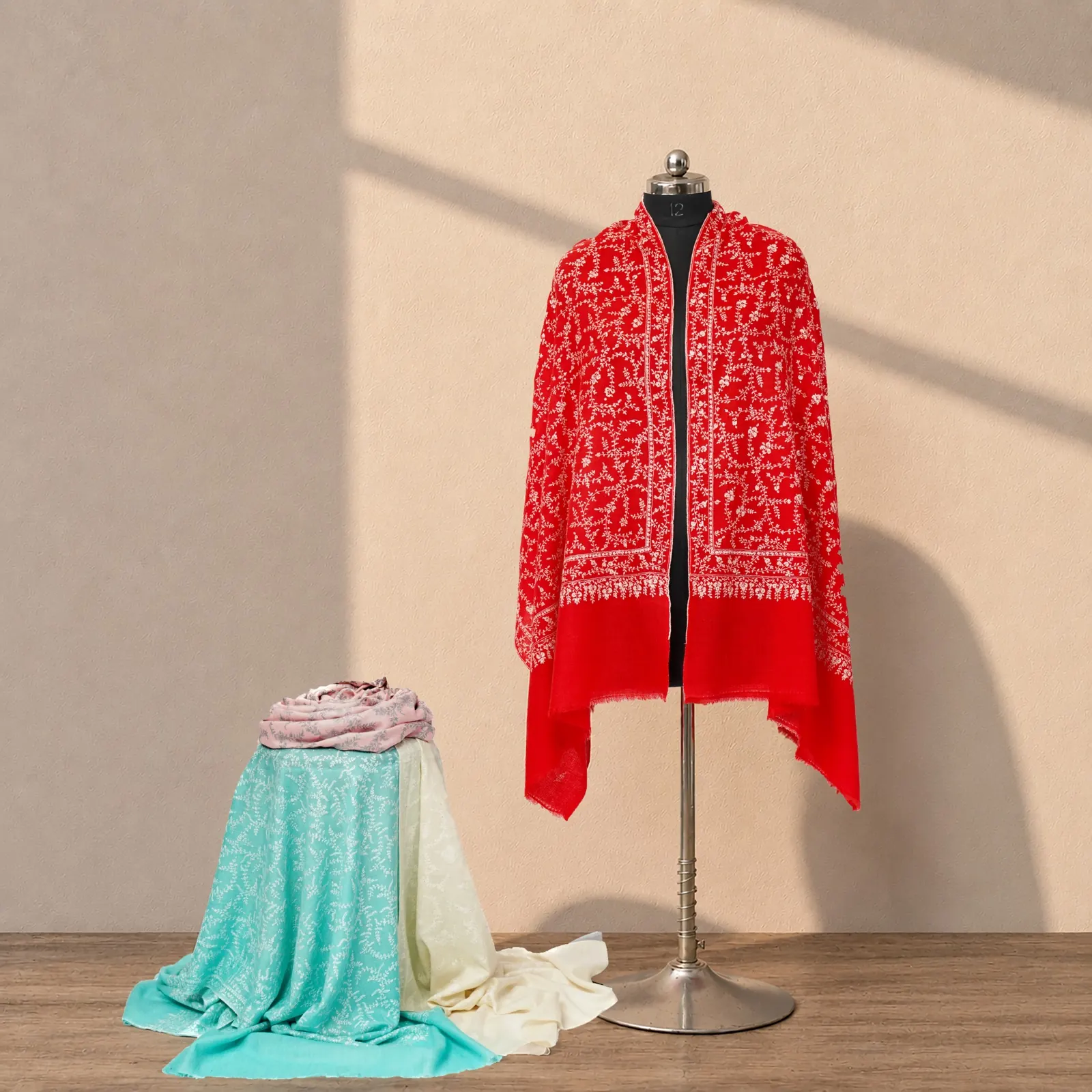 Clear red shawl covered in ivory needlework, a folded stack of pastel shawls on a table beside it.