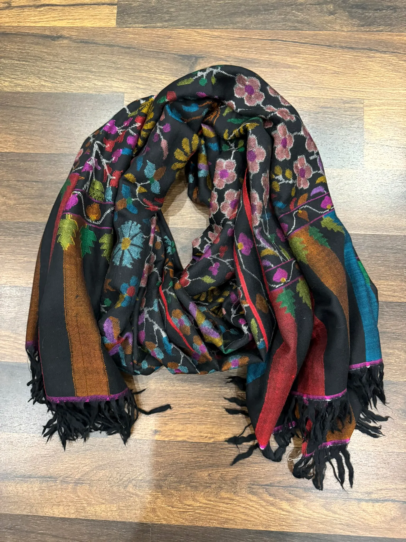 Black kani stole covered in multicoloured flowers, with a black fringe, on a wooden floor.