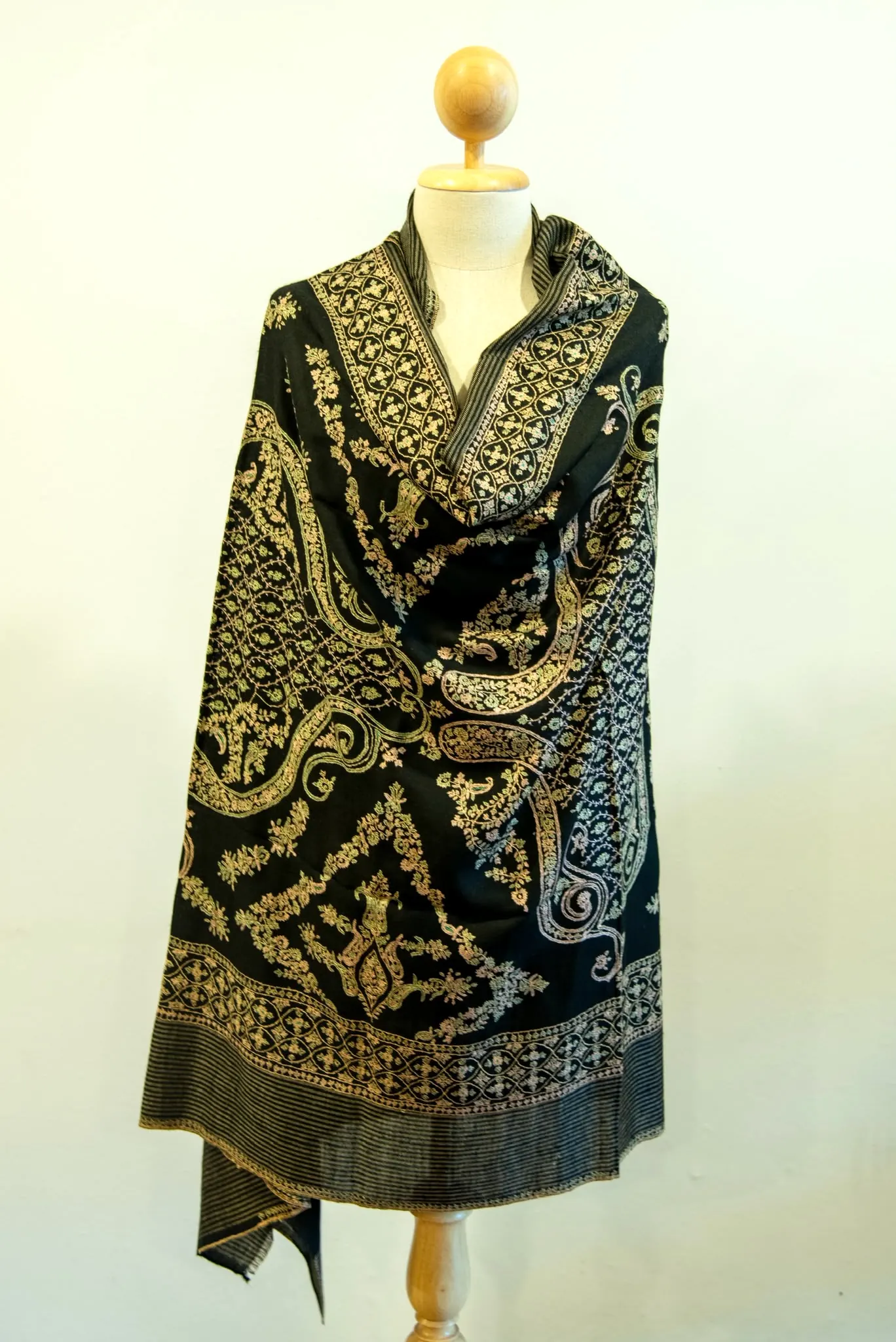 Black shawl covered in gold and ivory paisley needlework, draped on a stand.
