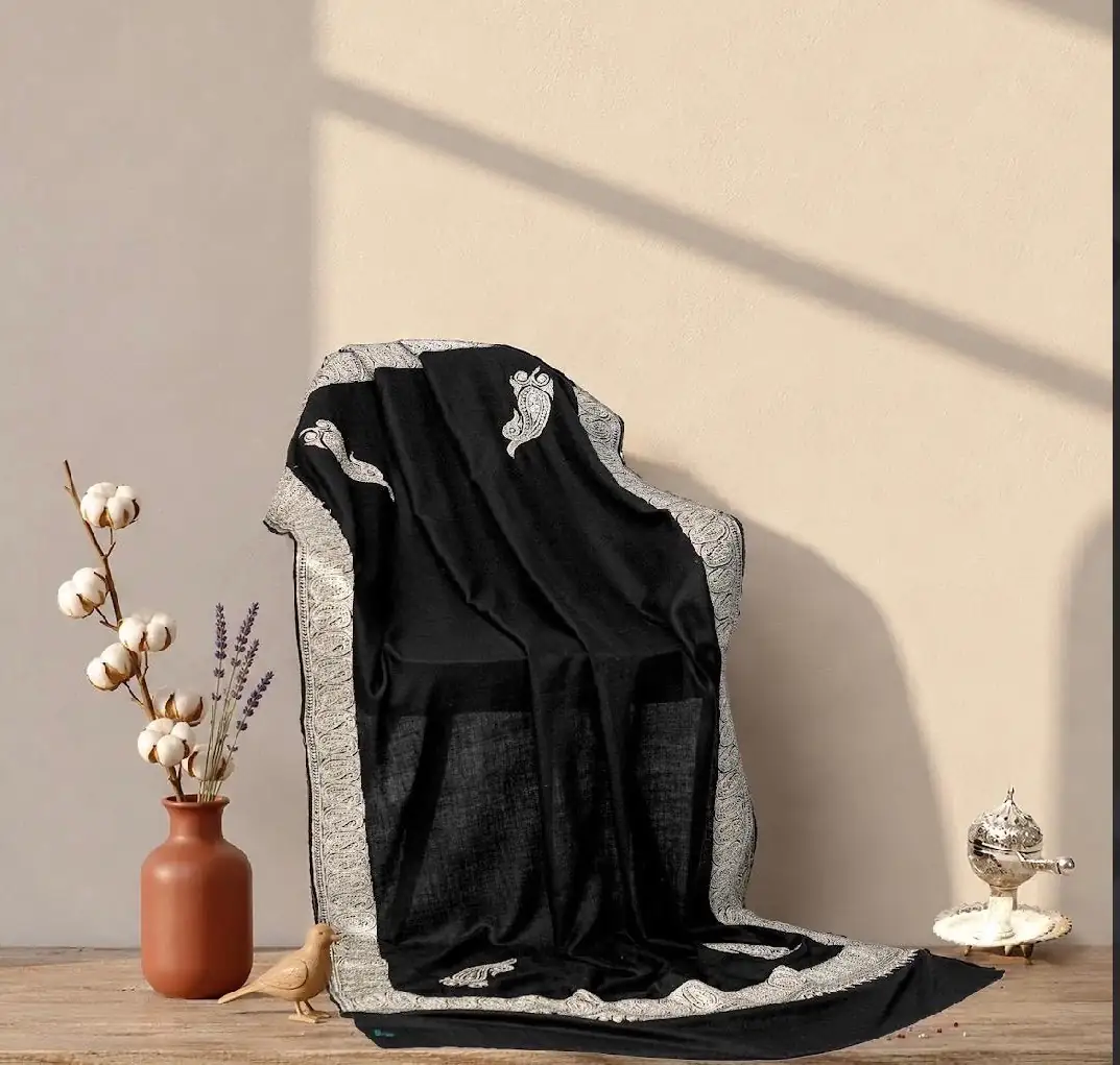 Black shawl with a broad silver metallic border and scattered motifs, draped over a chair beside a vase of cotton and lavender.