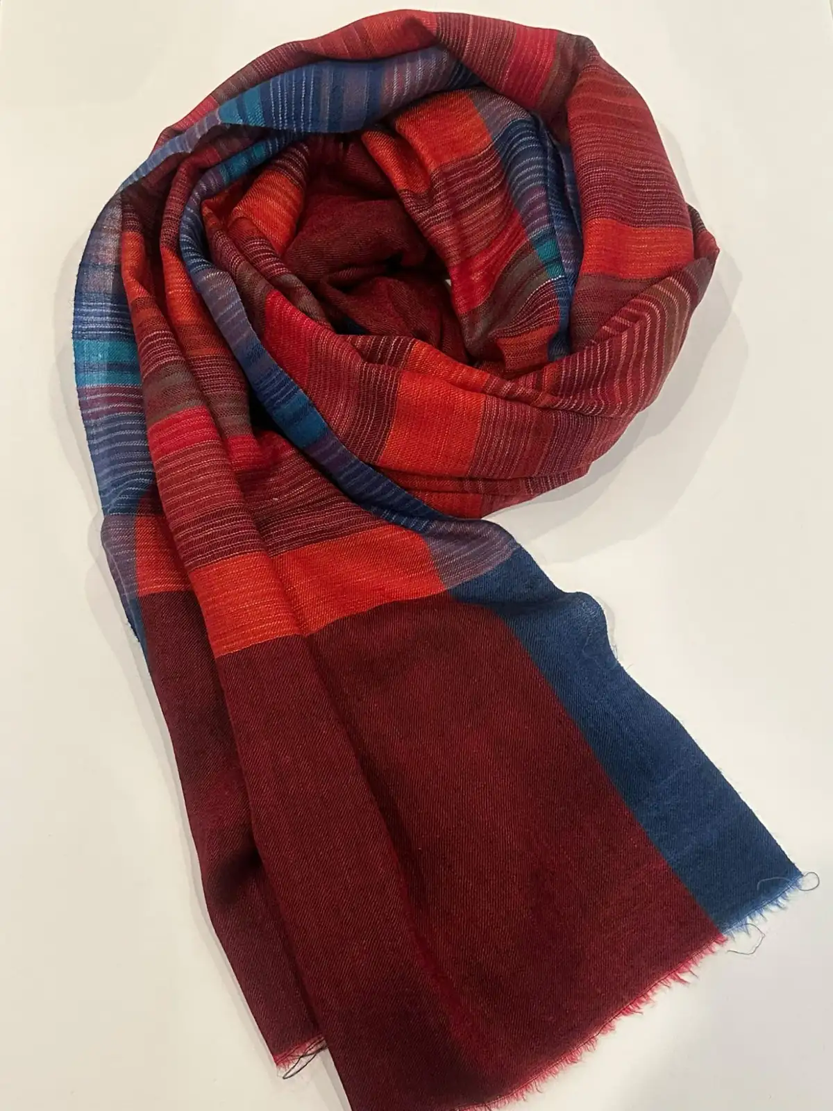 Woven stole in crimson, orange and blue checks and stripes, loosely coiled on a white surface.