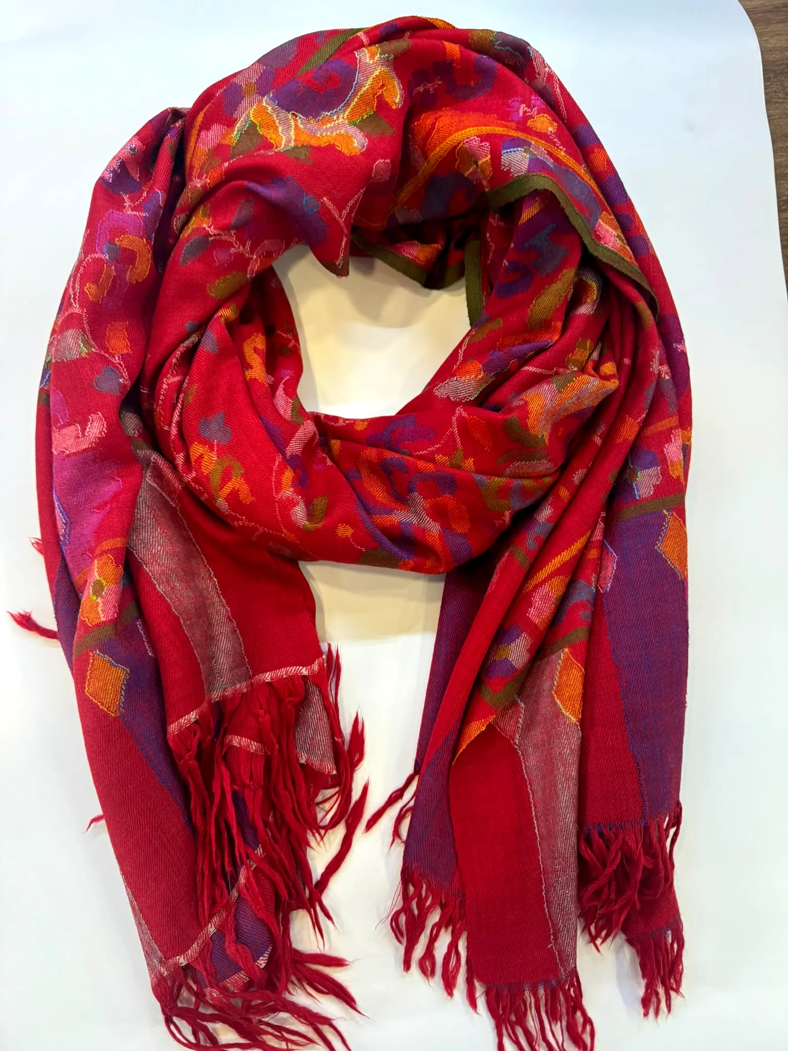 Crimson kani stole with pink, orange and violet florals and a long red fringe, laid on a white table.