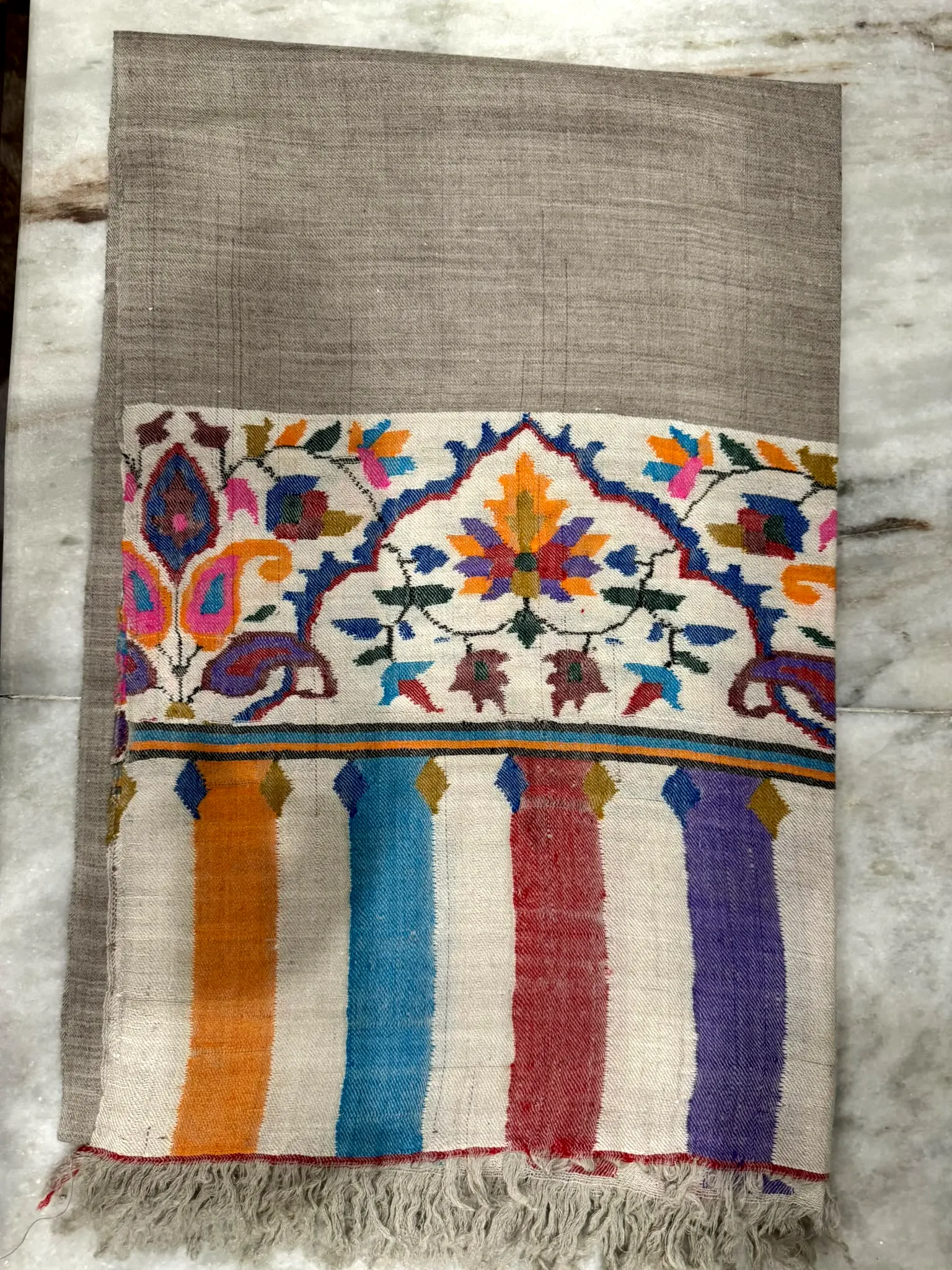 Grey kani shawl laid flat, showing an ivory border of paisley and flowers above bright vertical stripes.