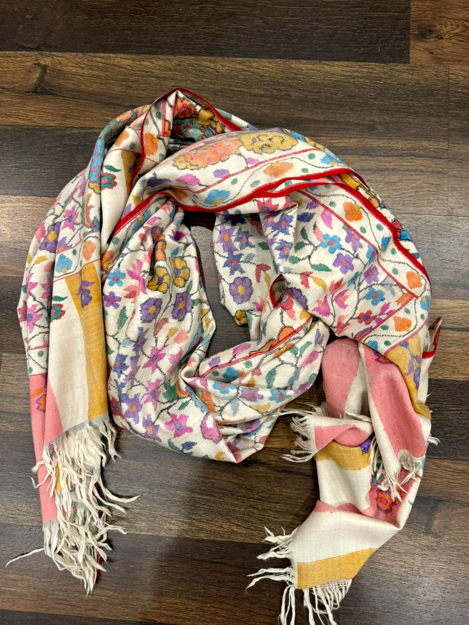 Ivory kani stole with multicoloured florals, a red edge and yellow and pink end panels, on a wooden floor.