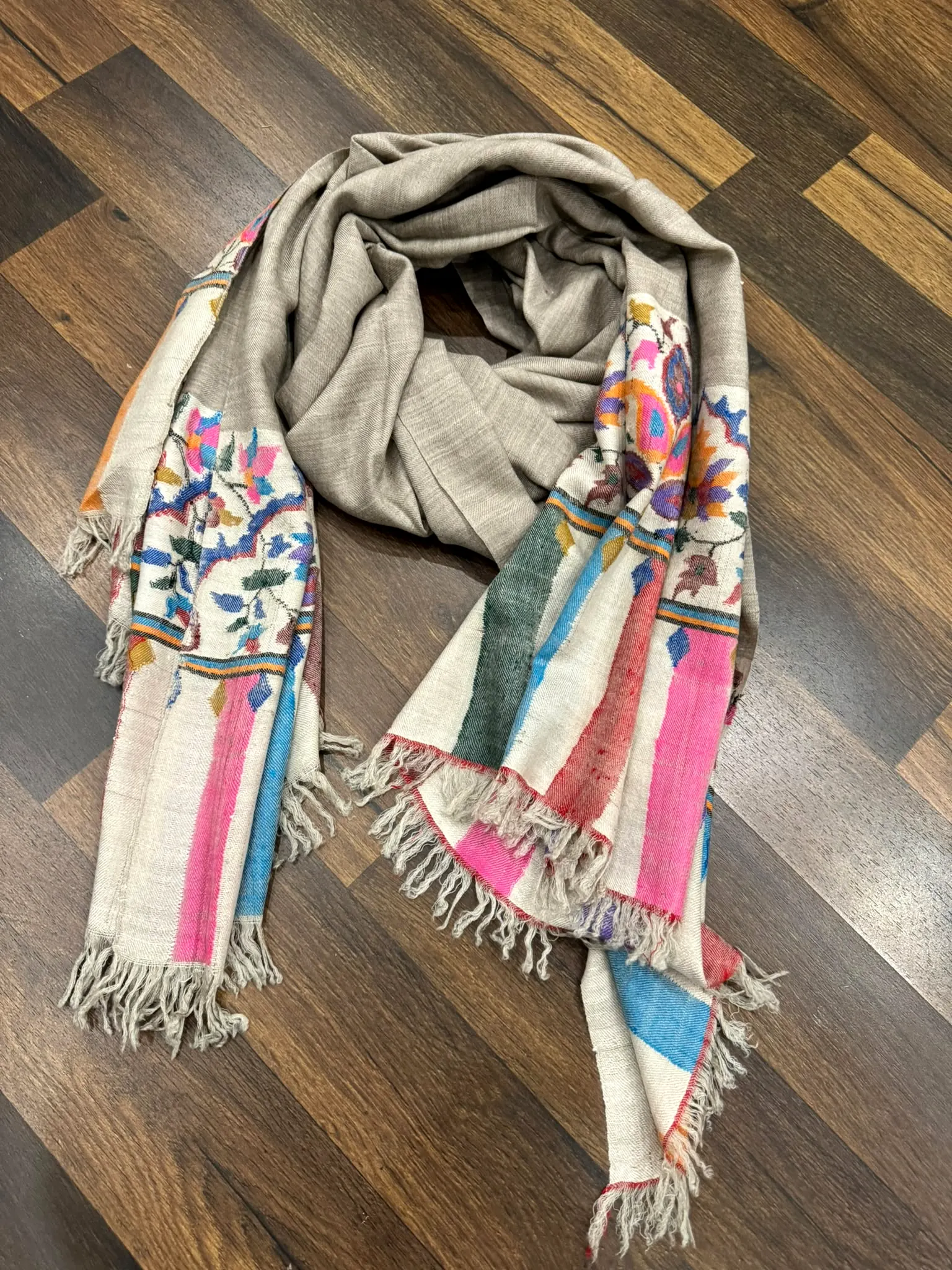 Oatmeal kani stole laid on a wooden floor, with pink, blue and floral striped ends.