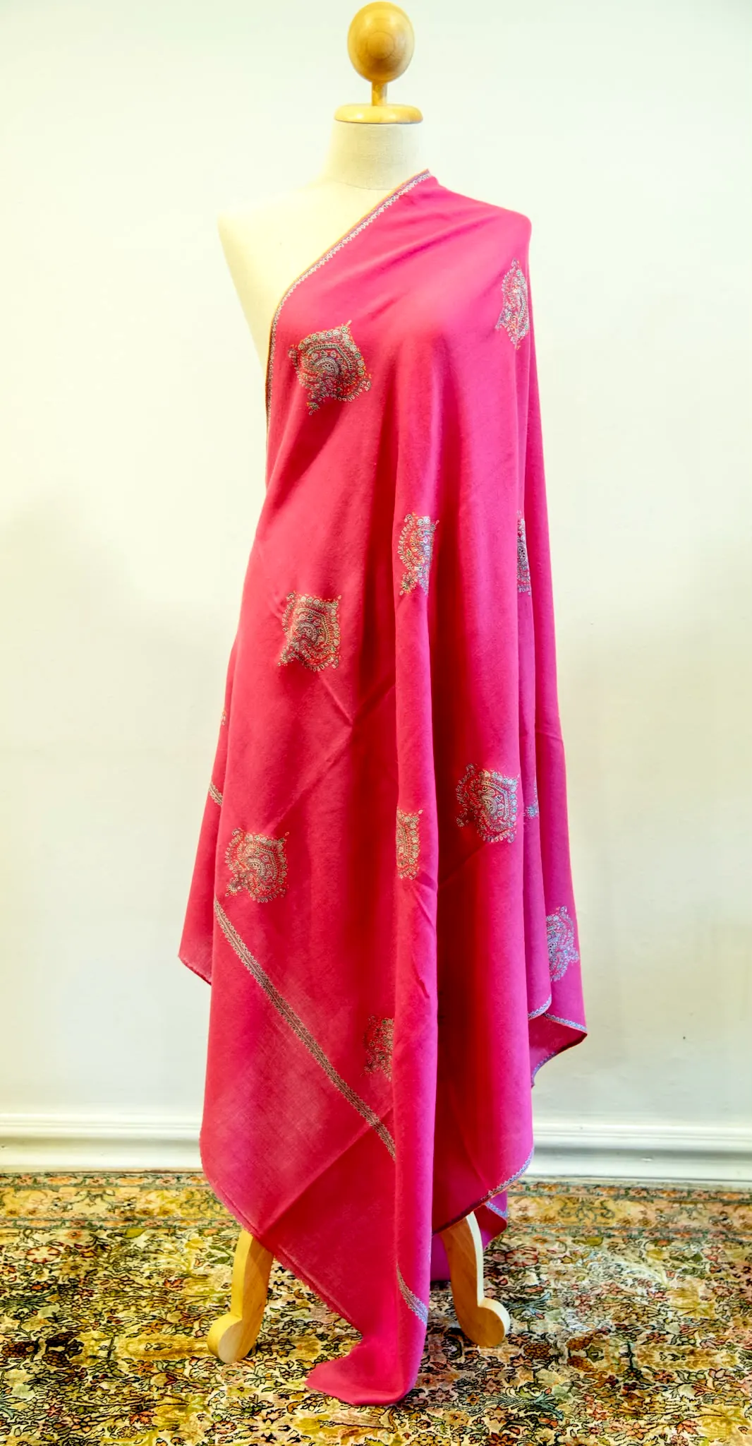 Hot pink shawl with gold toned needlework motifs, draped on a stand.