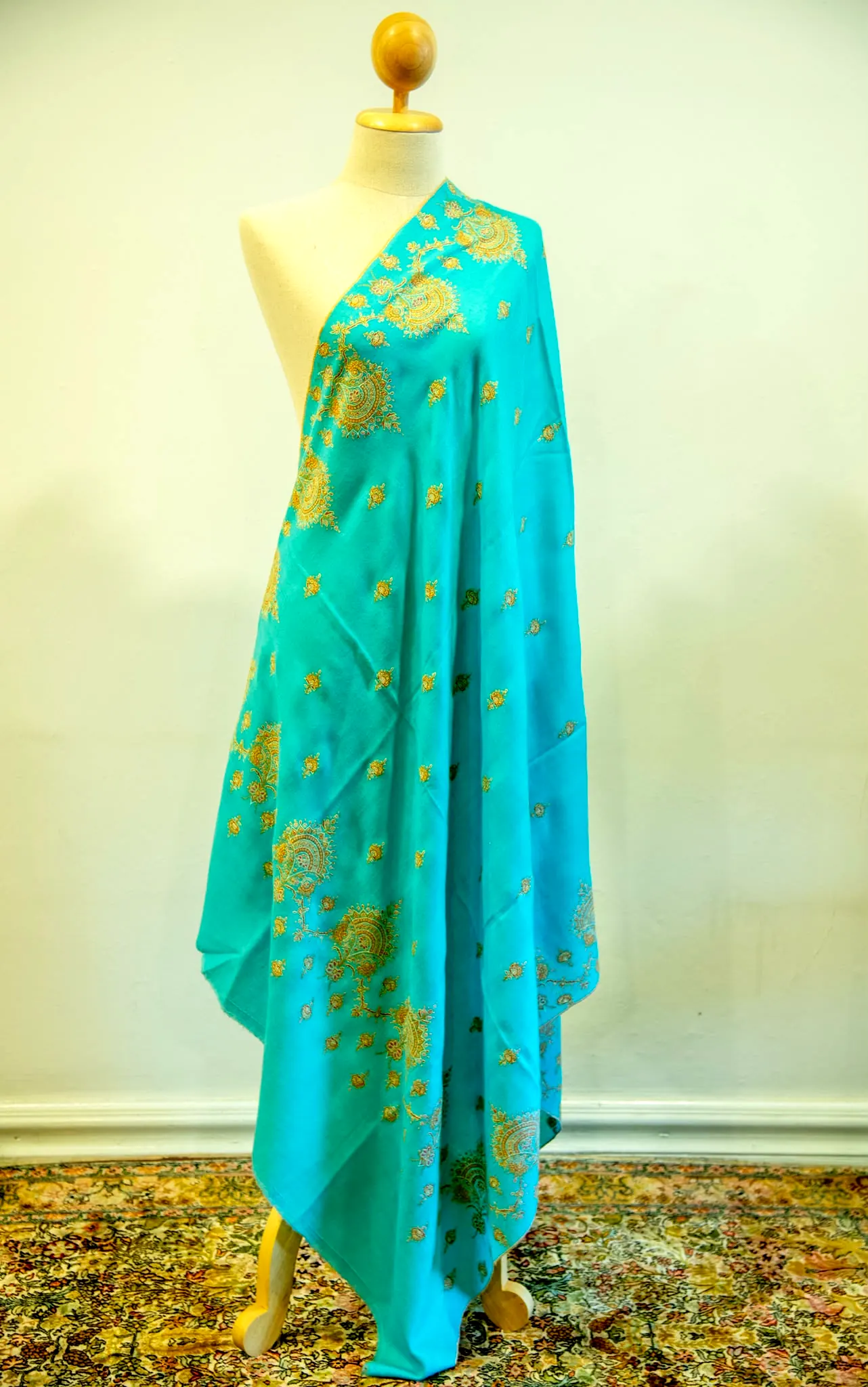 Turquoise shawl with gold toned needlework motifs, draped on a stand.
